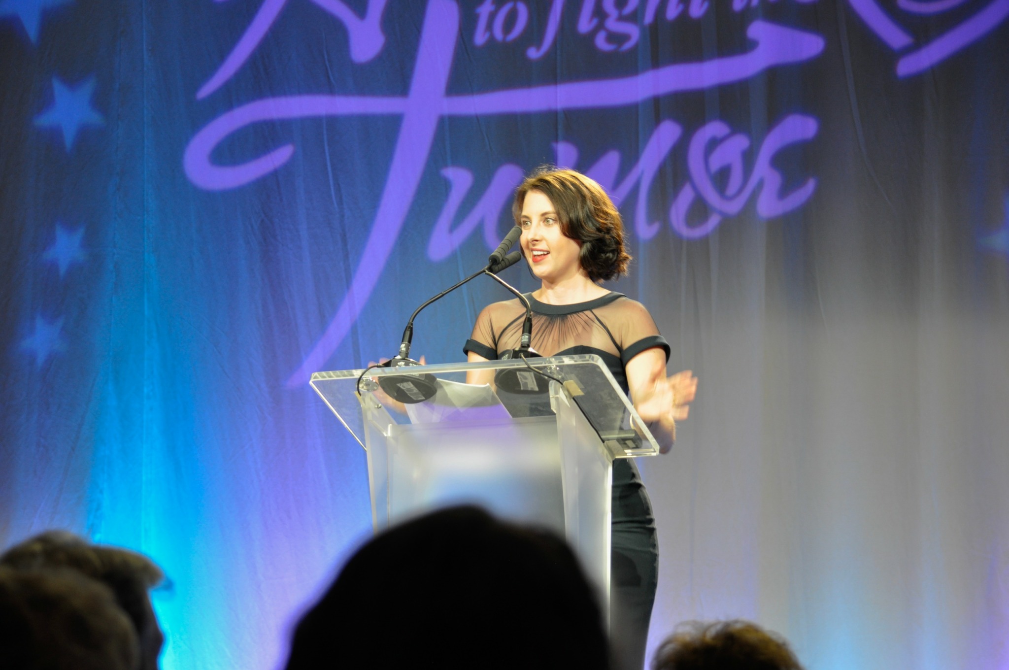 Woman speaking at a podium on stage with a purple and blue background, audience in foreground.
