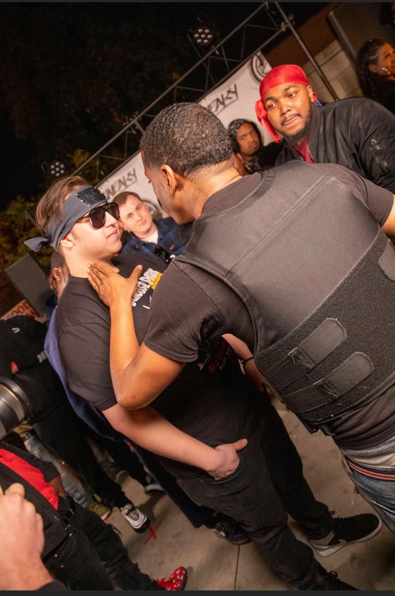 Police officer talking to woman with sunglasses, surrounded by people at night, some wearing red headbands, outdoor setting.
