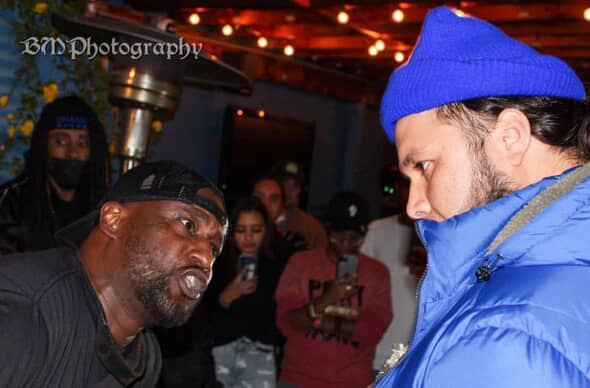 Two men face each other closely in a dimly lit setting with string lights overhead, surrounded by people. One wears a black cap, the other a blue beanie and jacket.