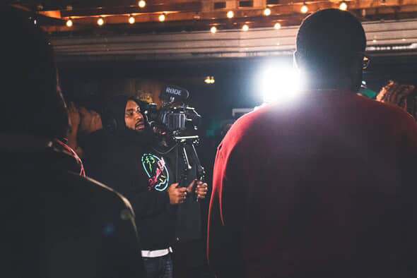 People at an indoor event with a bright light in the background, some holding cameras, dim lighting, and a ceiling with string lights.