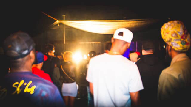 People gathered at an outdoor event at night, facing a bright stage or screen, with some wearing hats and casual clothing.