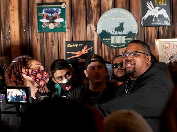 Group of people gathered indoors, smiling and talking, with wooden wall decorated with holiday-themed signs in the background.