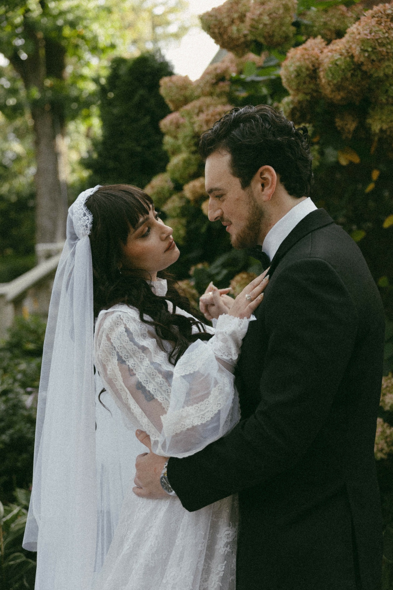 A bride and groom face each other outdoors, surrounded by greenery and flowers, holding hands and smiling.