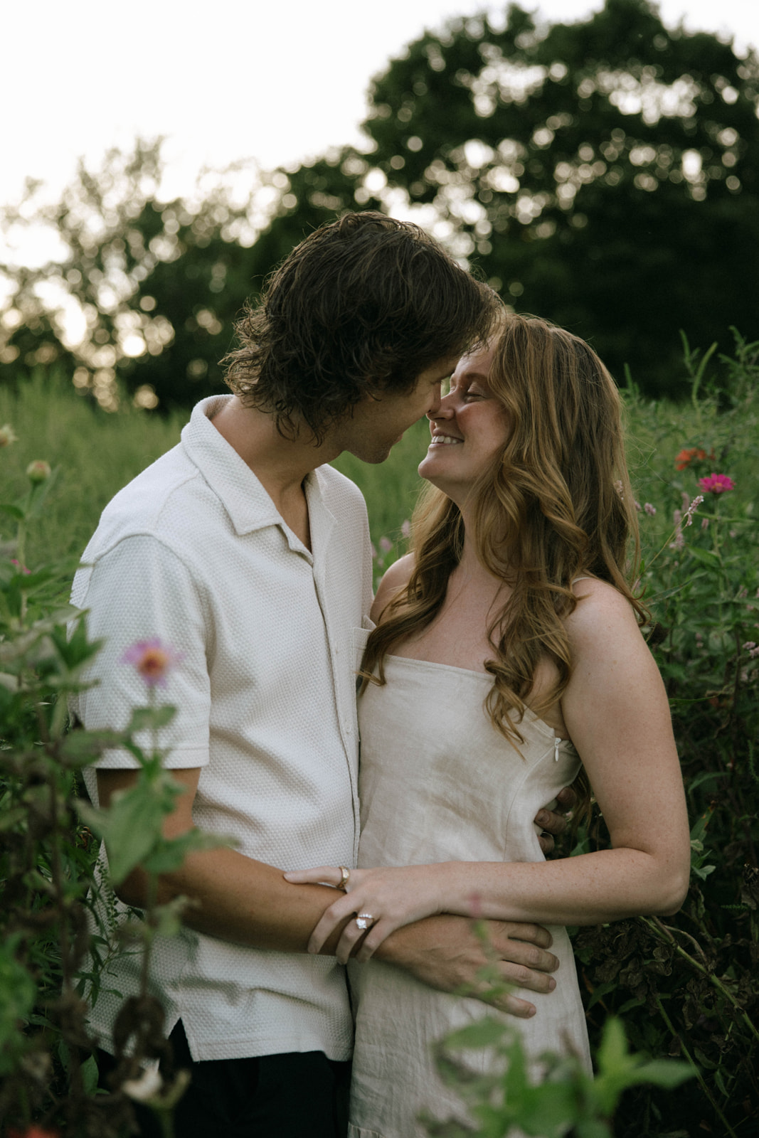A couple stands close together outdoors, smiling and looking at each other, surrounded by greenery and flowers.