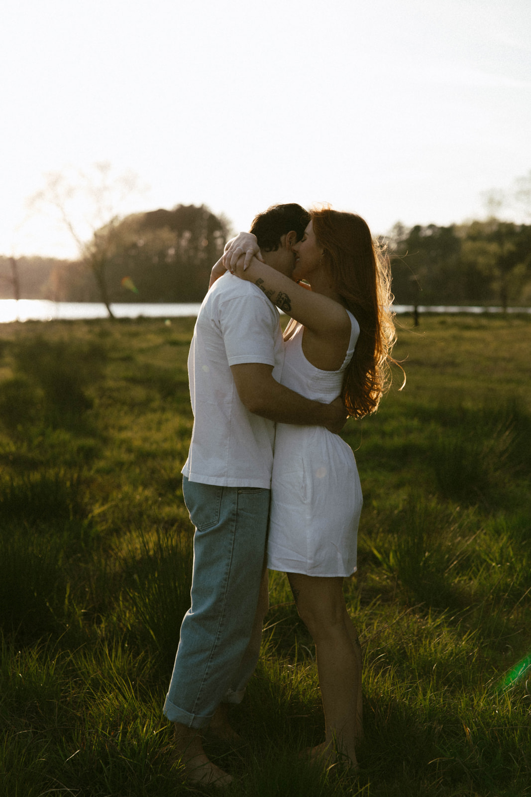 A couple embraces outdoors during sunset, standing on grass with trees and water in the background.