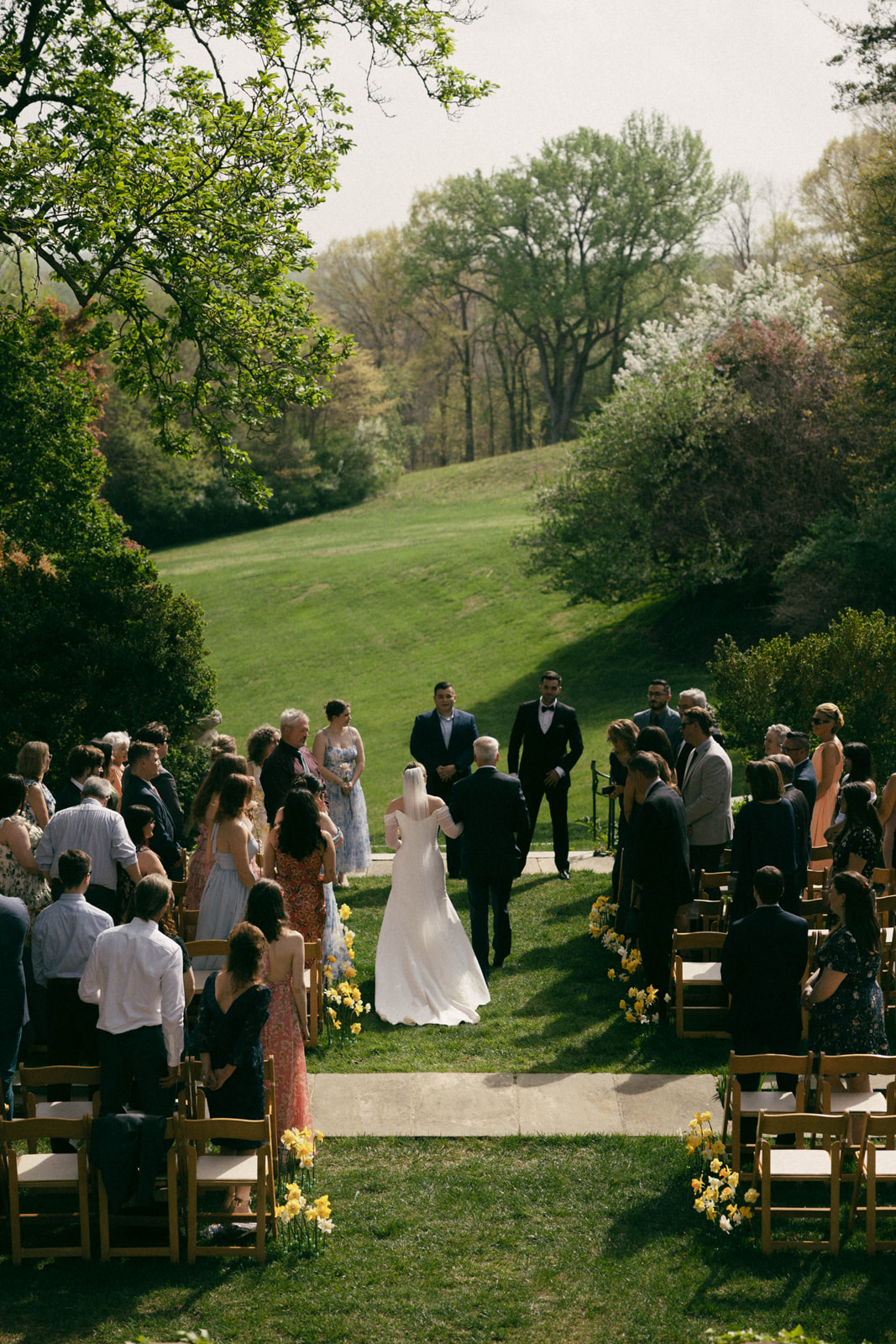 Wedding ceremony outdoors with guests seated, bride and groom standing, surrounded by trees and greenery.