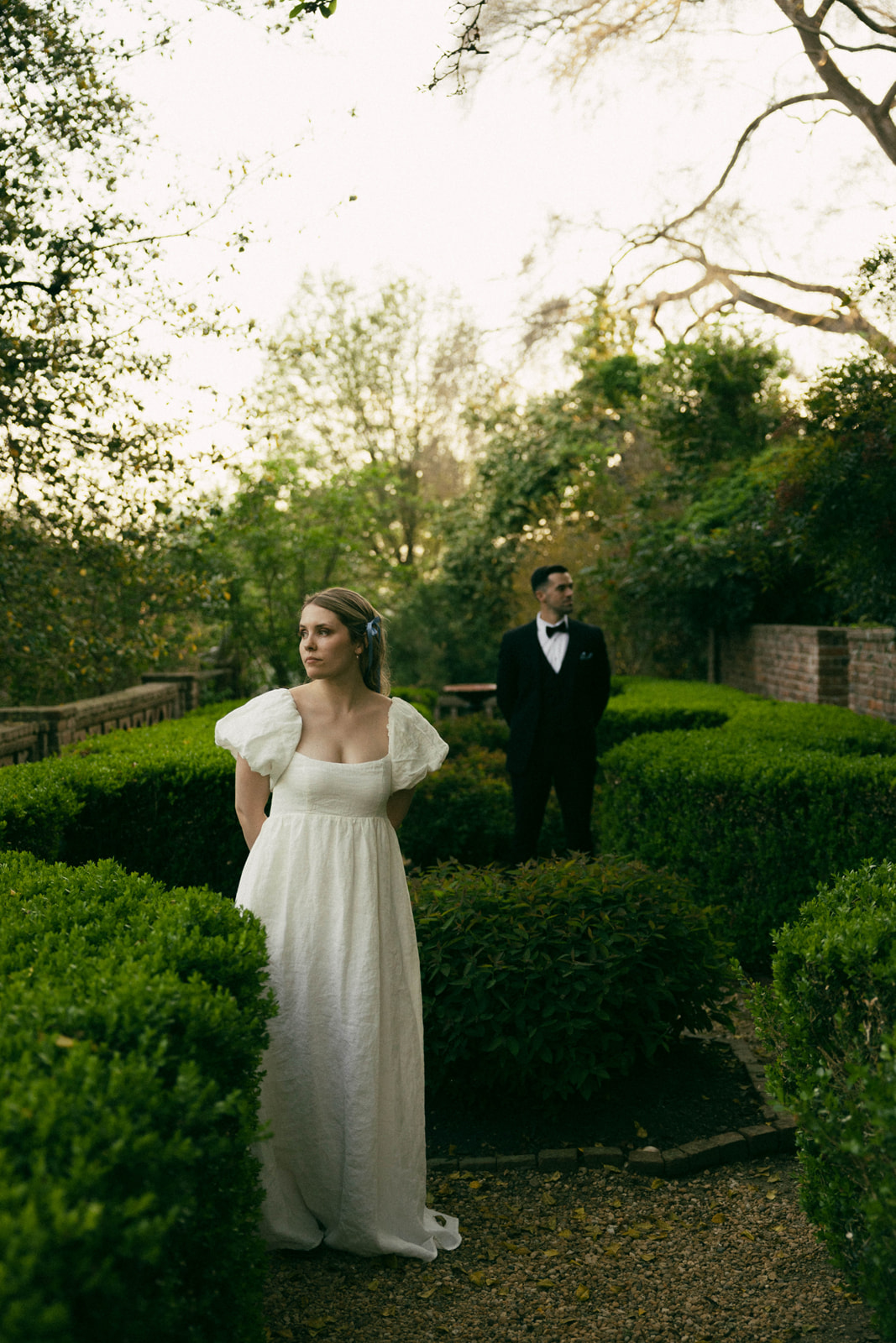 A woman in a white dress stands in a garden with greenery, while a man in a tuxedo stands behind her.