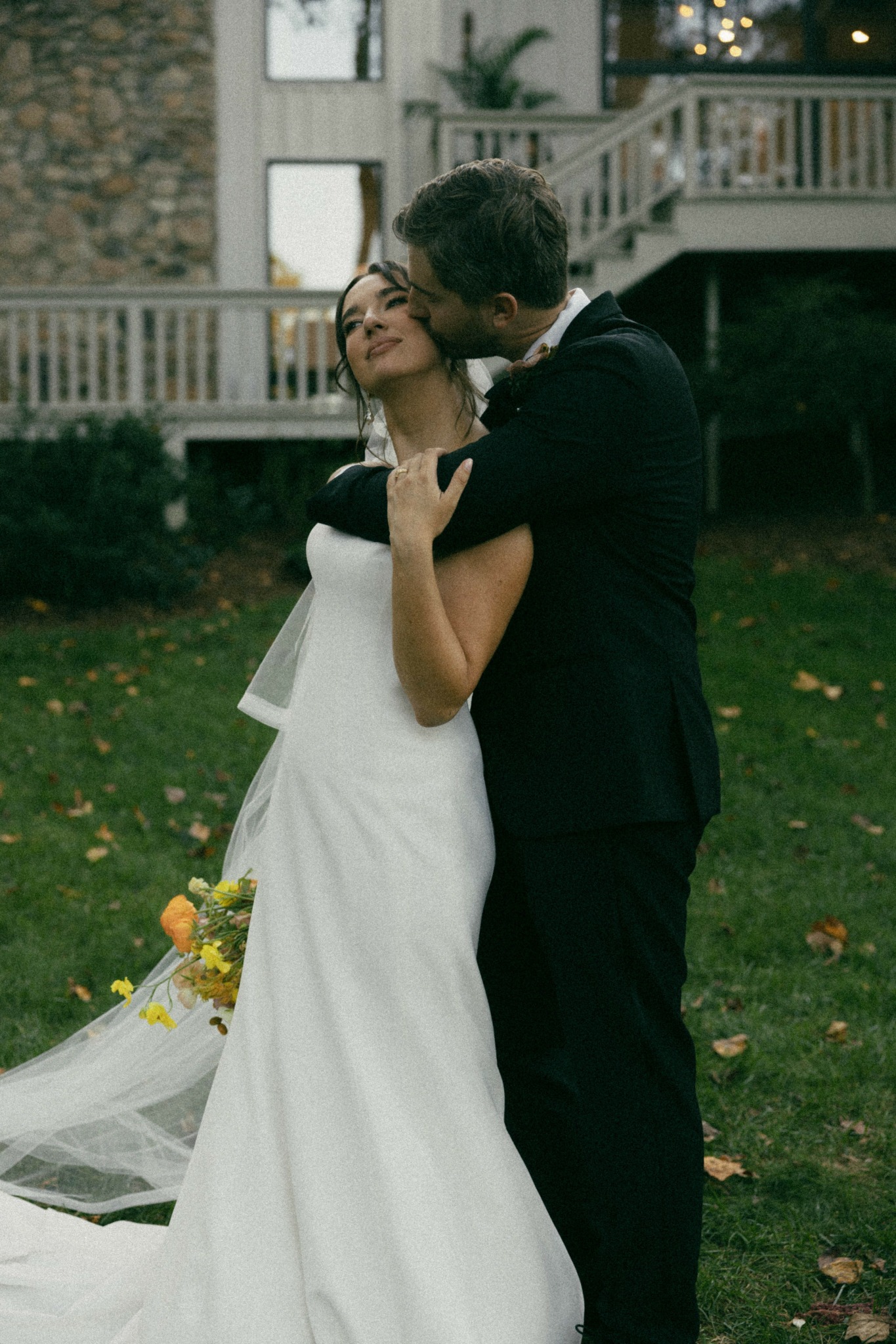 A couple in wedding attire embracing outdoors, with a house and greenery in the background.