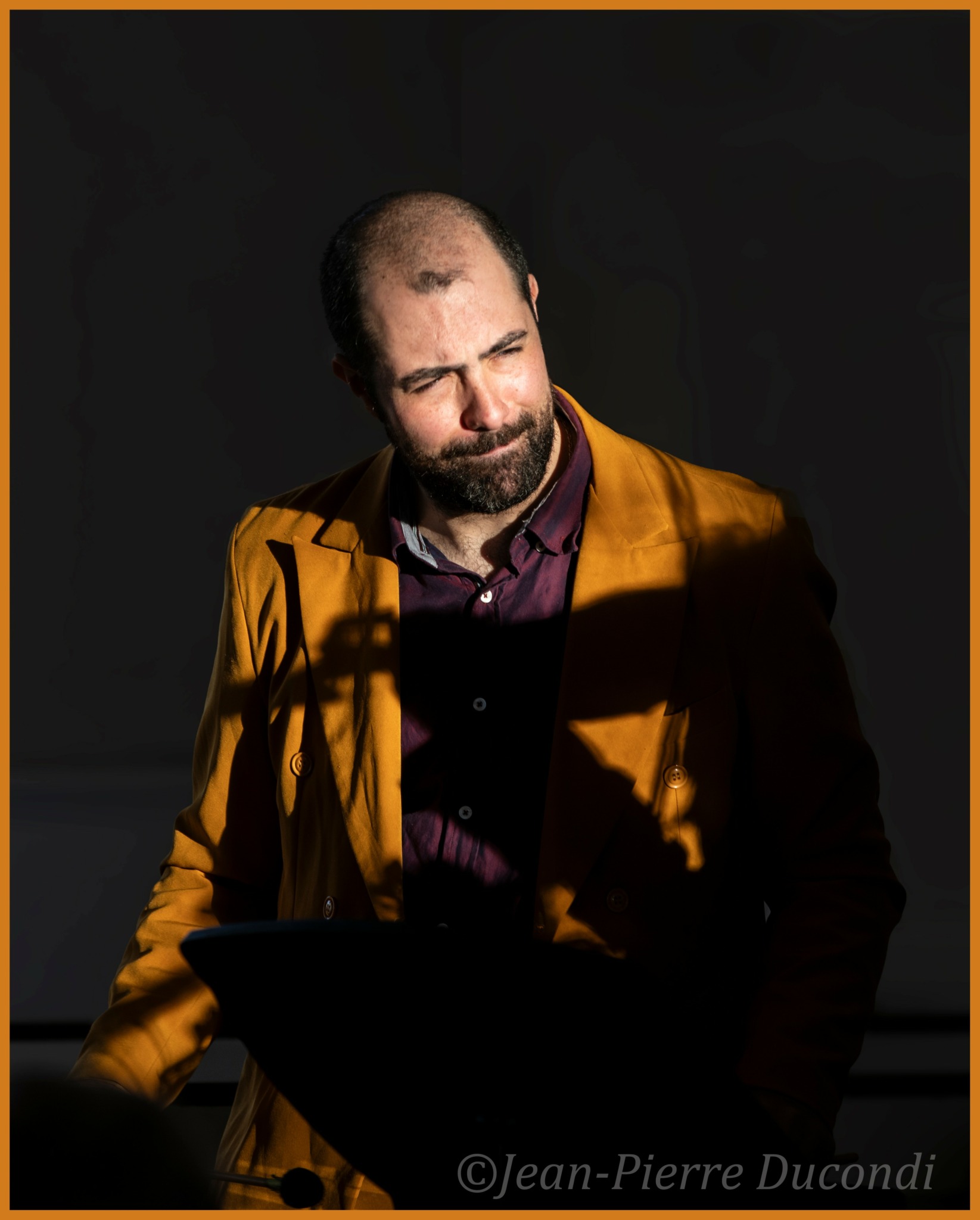 Man with beard in yellow jacket and dark shirt, looking down, shadow cast on face and background, dark background