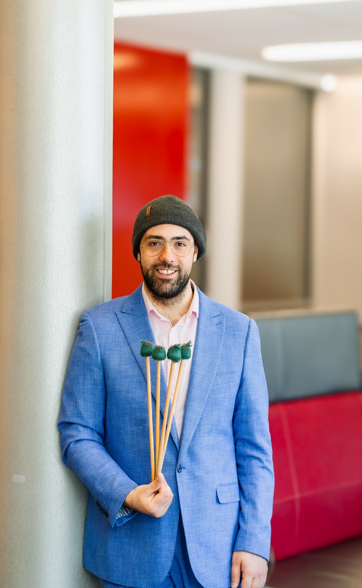 Man with a beard wearing a beanie and blue blazer, holding green lollipops, standing indoors near a wall.