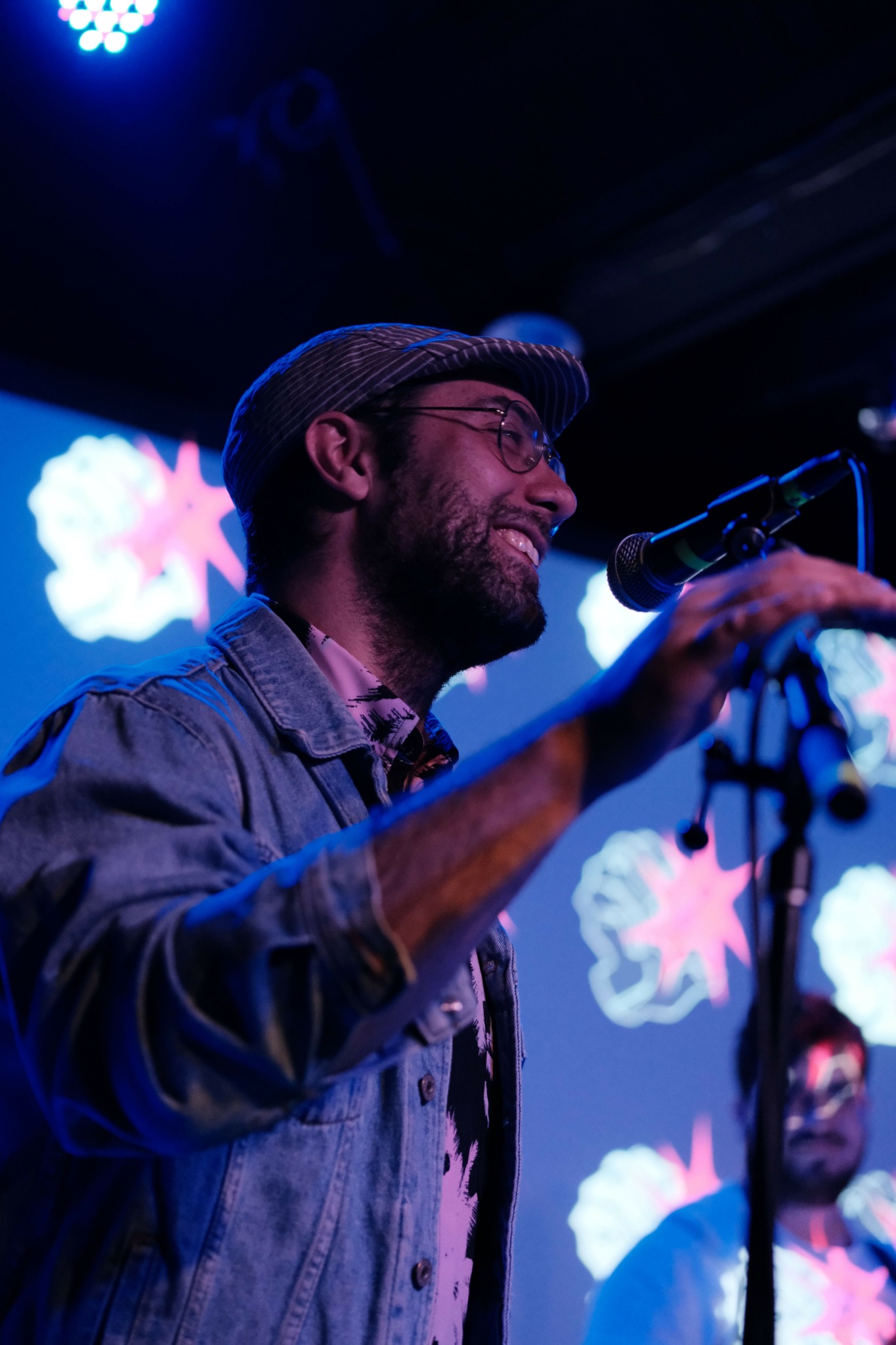 A man wearing glasses and a cap plays a microphone on stage with colorful lights and patterns in the background.