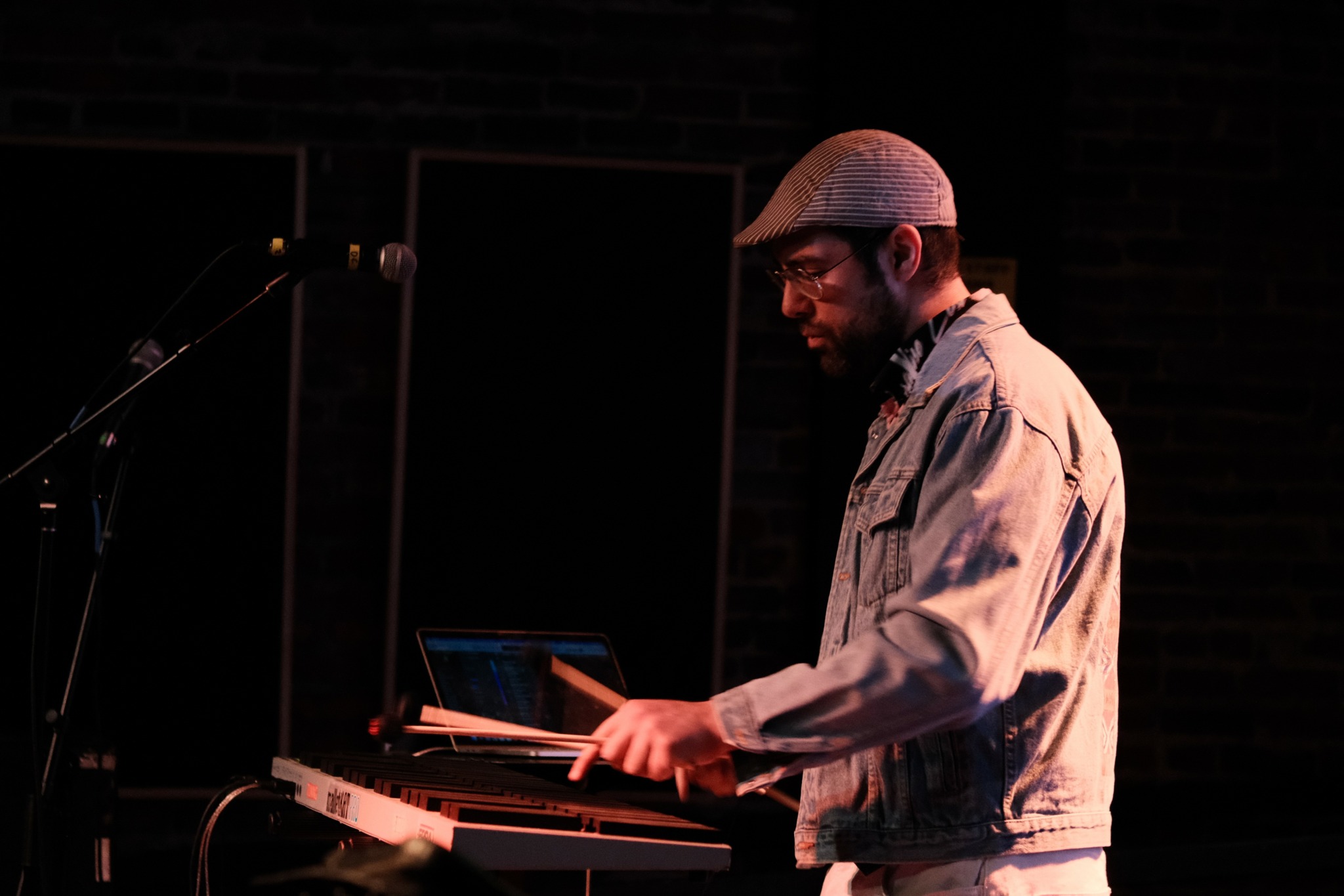 Person wearing a hat and denim jacket playing keyboard on stage with dark background.