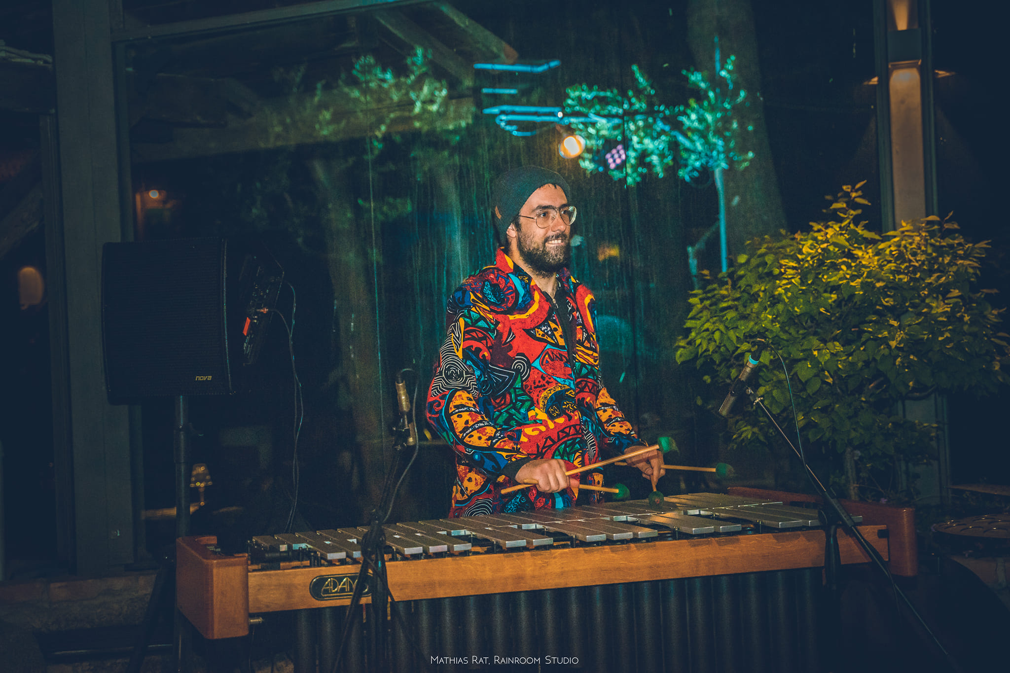Man with glasses and beard playing a percussion instrument at an indoor event, surrounded by plants and colorful lighting.
