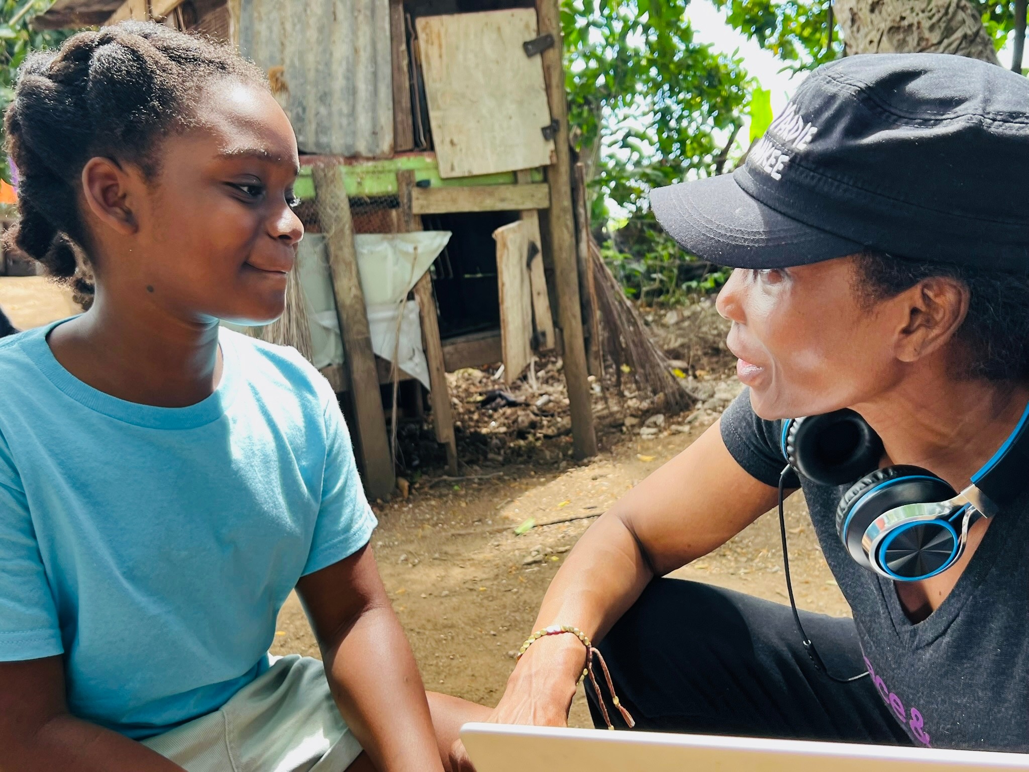 Two people, a girl and a woman, face each other outdoors, engaging in conversation with trees and a wooden structure in the background.