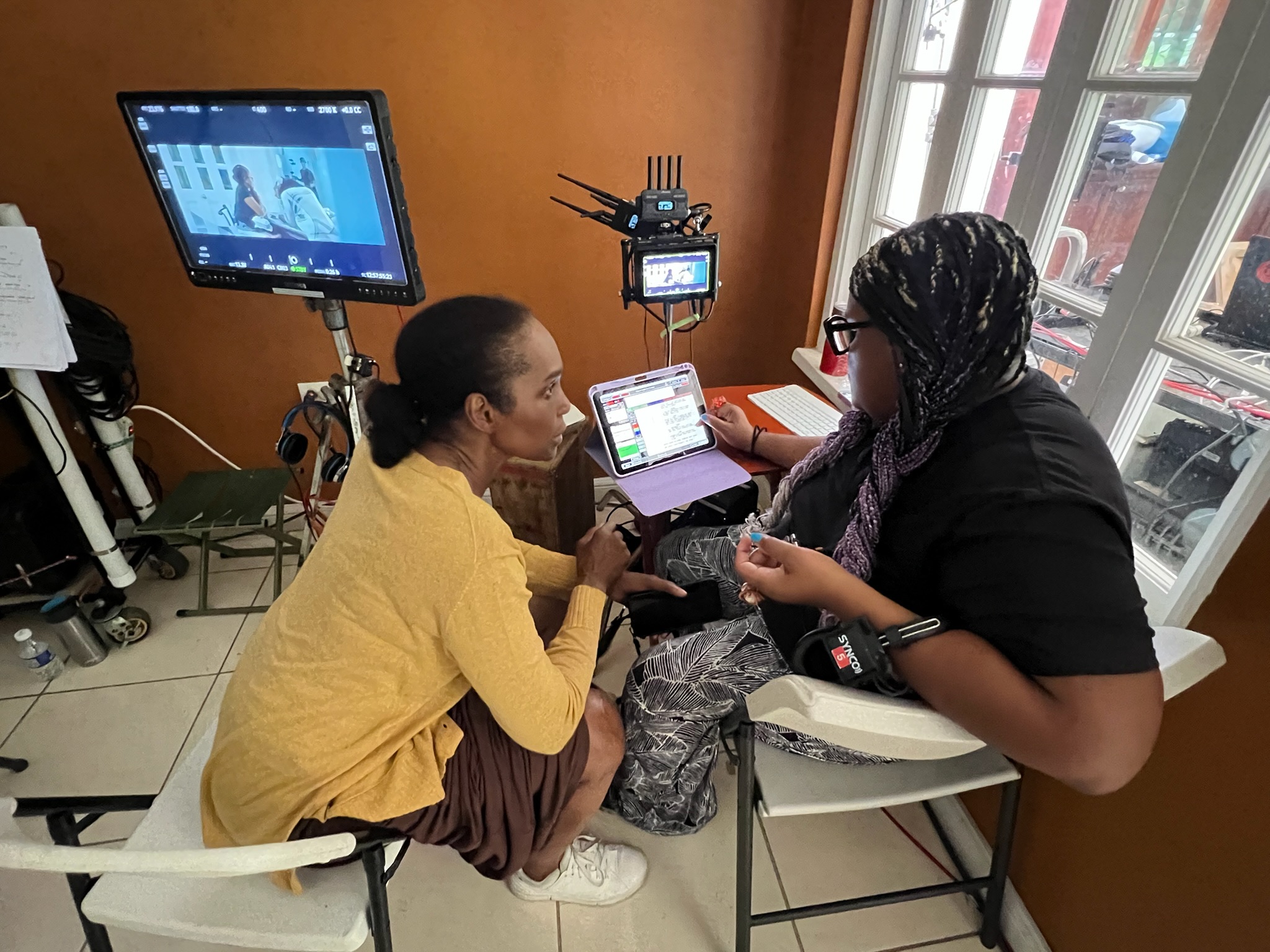 Two women sit at a table in a room with a window, engaged in conversation, with a monitor and camera equipment nearby.