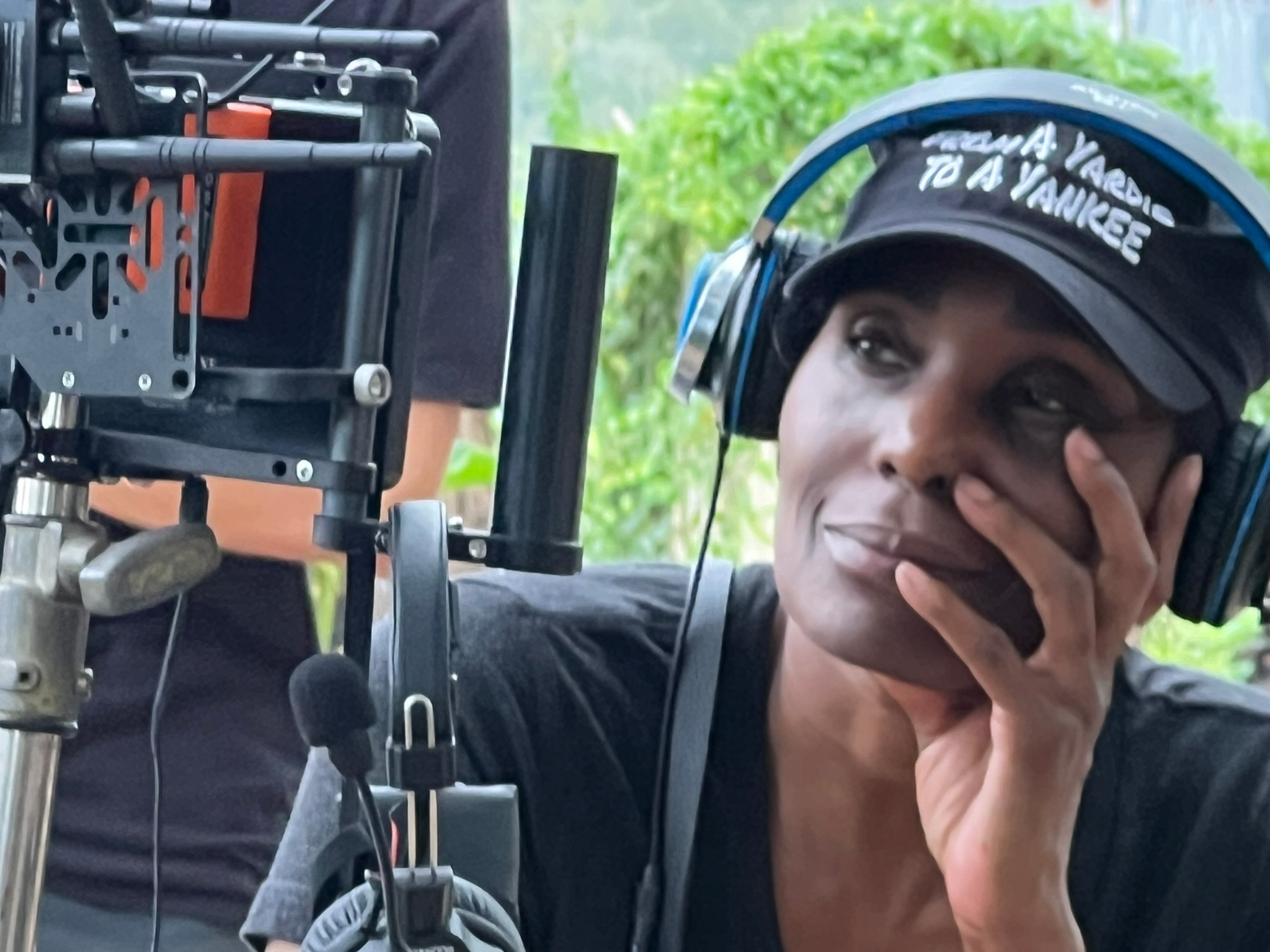 Woman wearing headphones and a cap, resting her face on her hand, outdoors with greenery in the background.