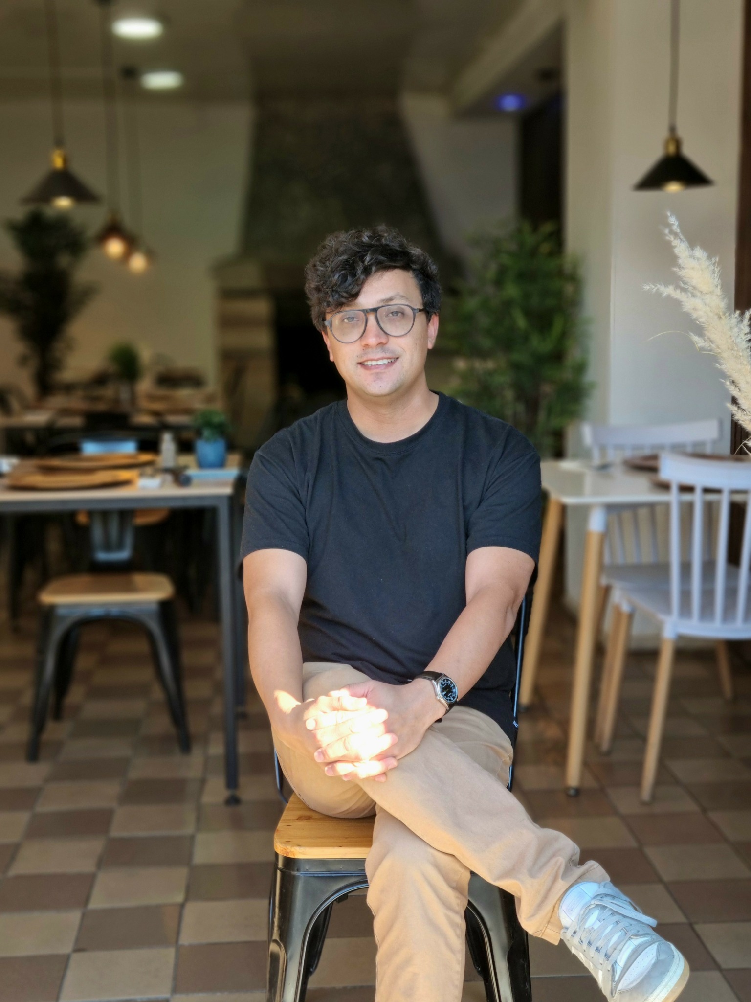 Young man with glasses sitting on a stool in a modern cafe, smiling, with tables and chairs in the background.