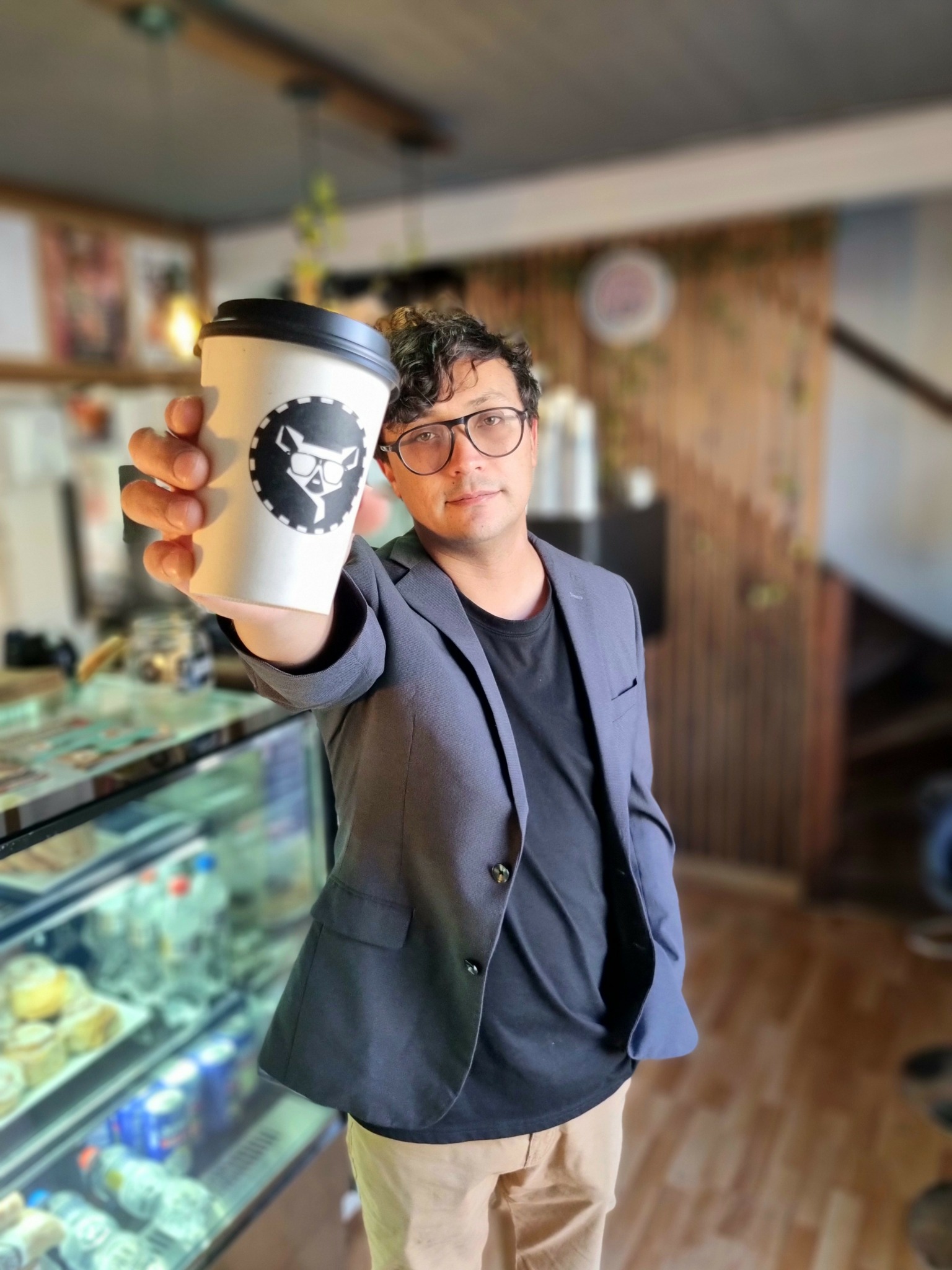 Person with glasses holding a coffee cup towards the camera inside a cafe, background with wooden walls and display case.