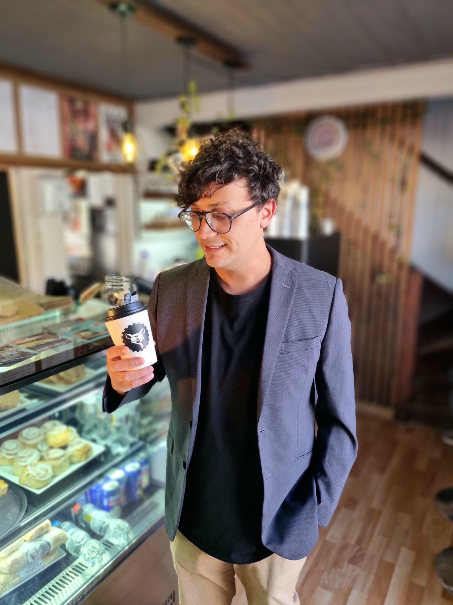 Young man with curly hair and glasses holding a coffee cup in a cafe, wearing a blazer and black shirt.