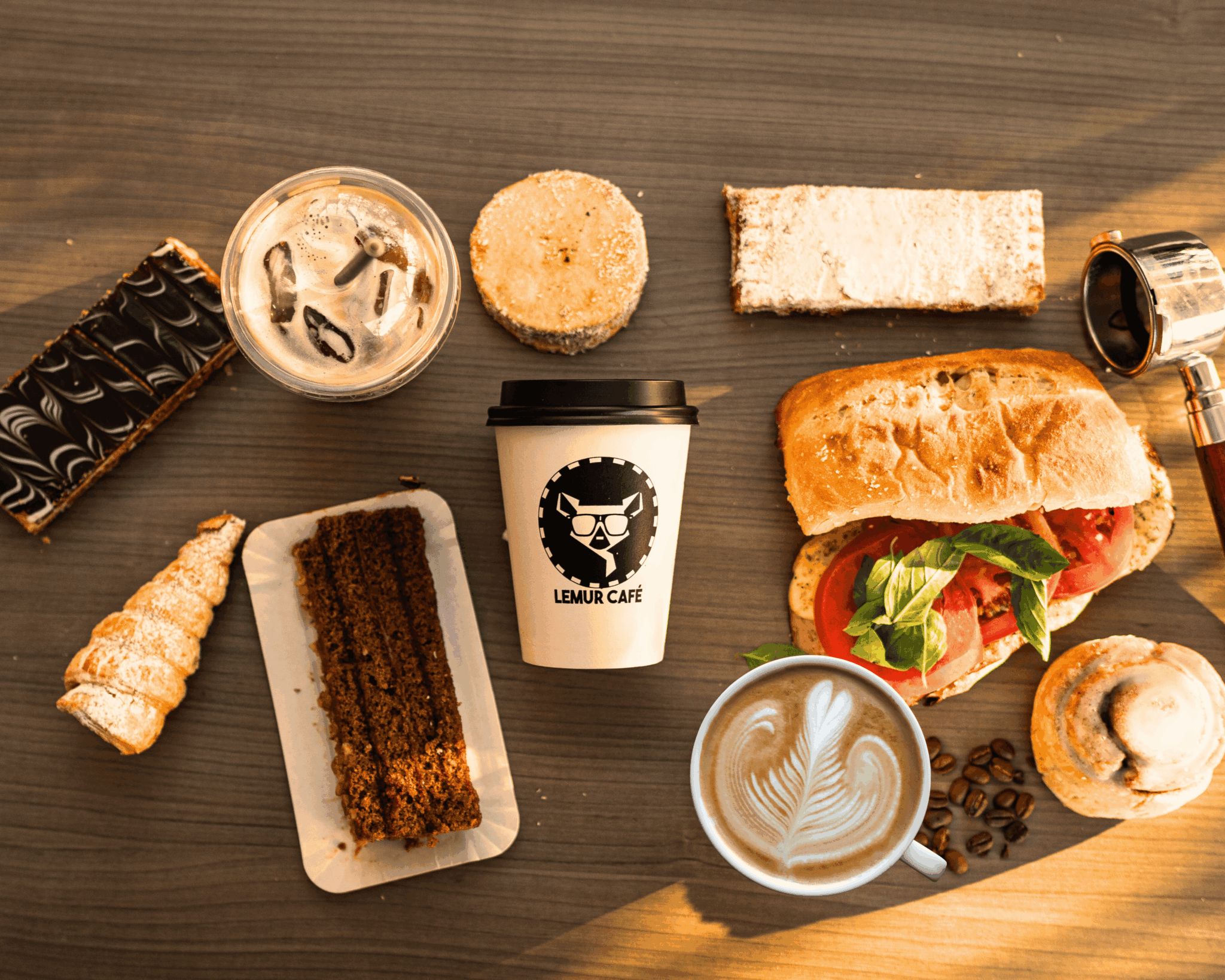 Assorted desserts, a sandwich with lettuce and tomato, a coffee cup, and a pastry on a wooden table.