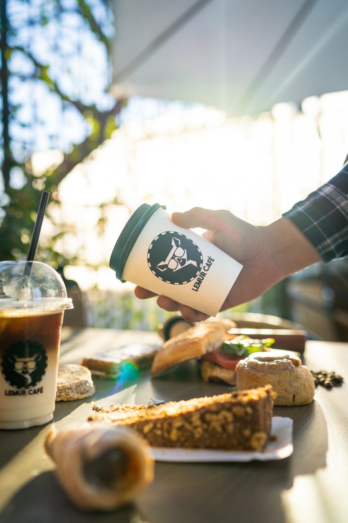 Person holding a coffee cup with a logo, surrounded by desserts on a table, sunlight streaming in through windows.
