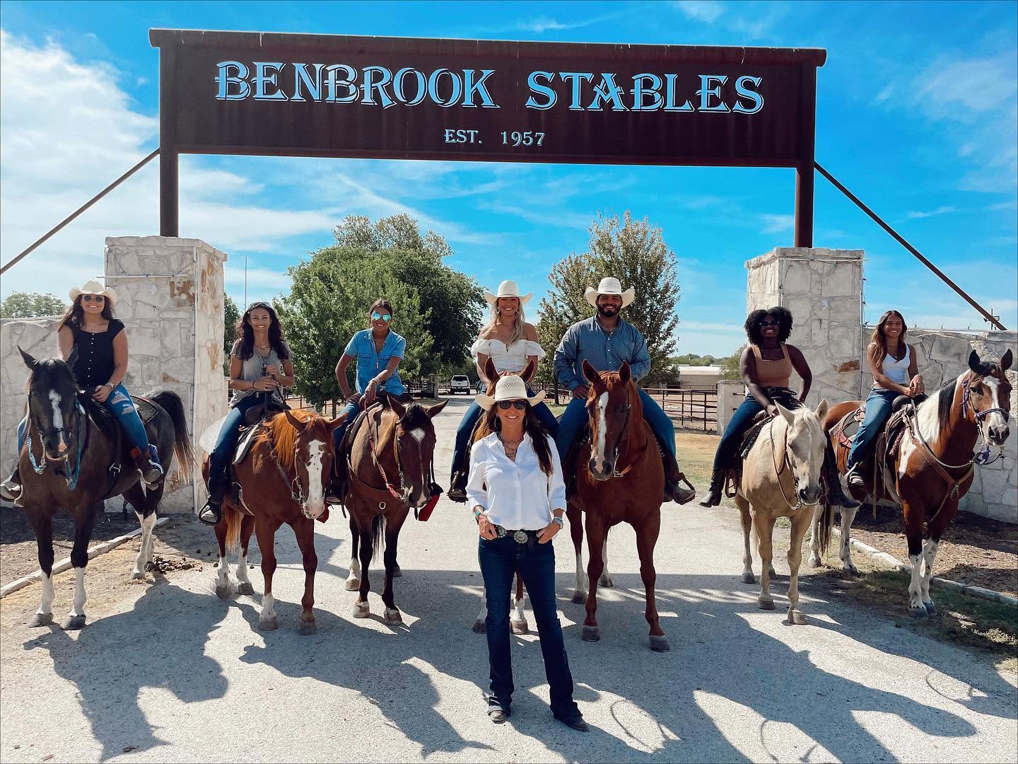 Group of people riding horses under a sign that reads 'Benbrook Stables' with trees and blue sky in background.