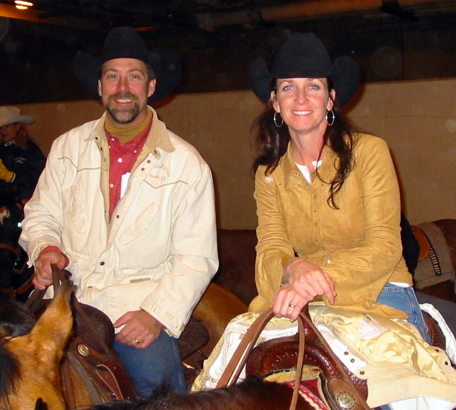 A man and woman in cowboy hats and western attire sit on horses indoors, smiling at the camera.