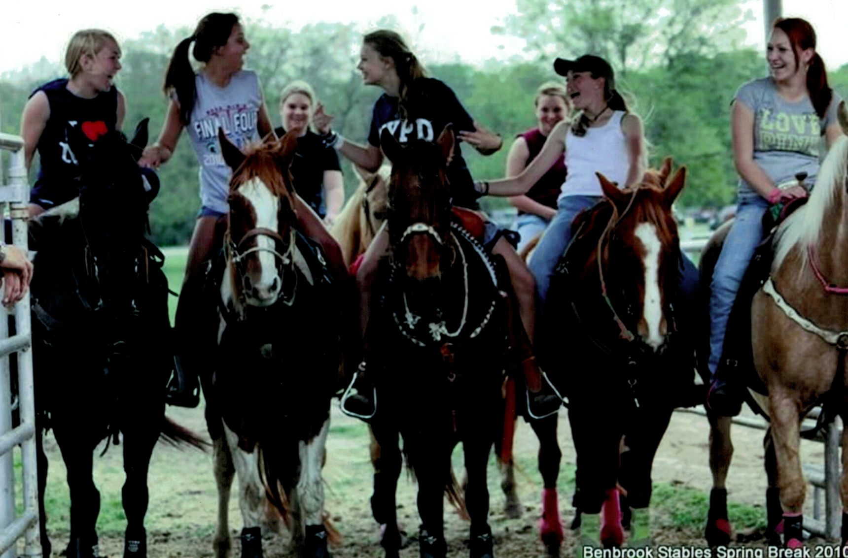Group of young women riding horses outdoors, some talking and smiling, with trees in the background.