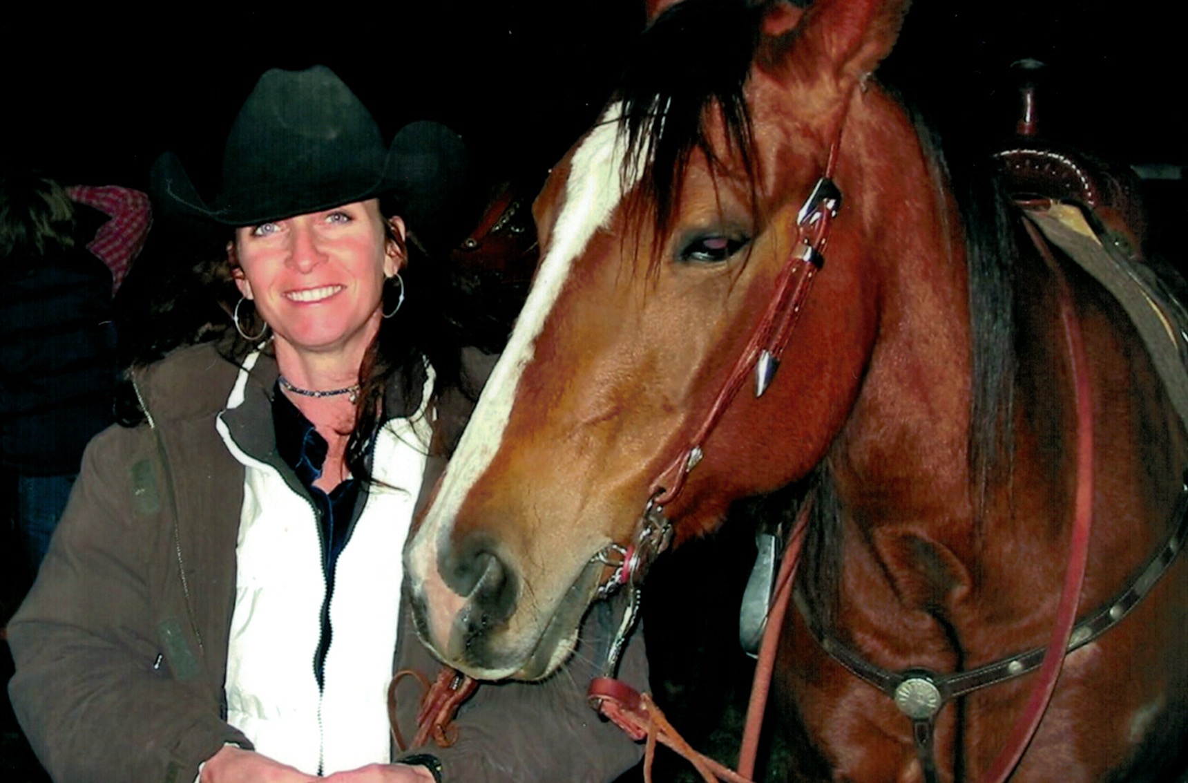 Woman with dark hair and hat standing next to a brown horse with a white stripe on its face.