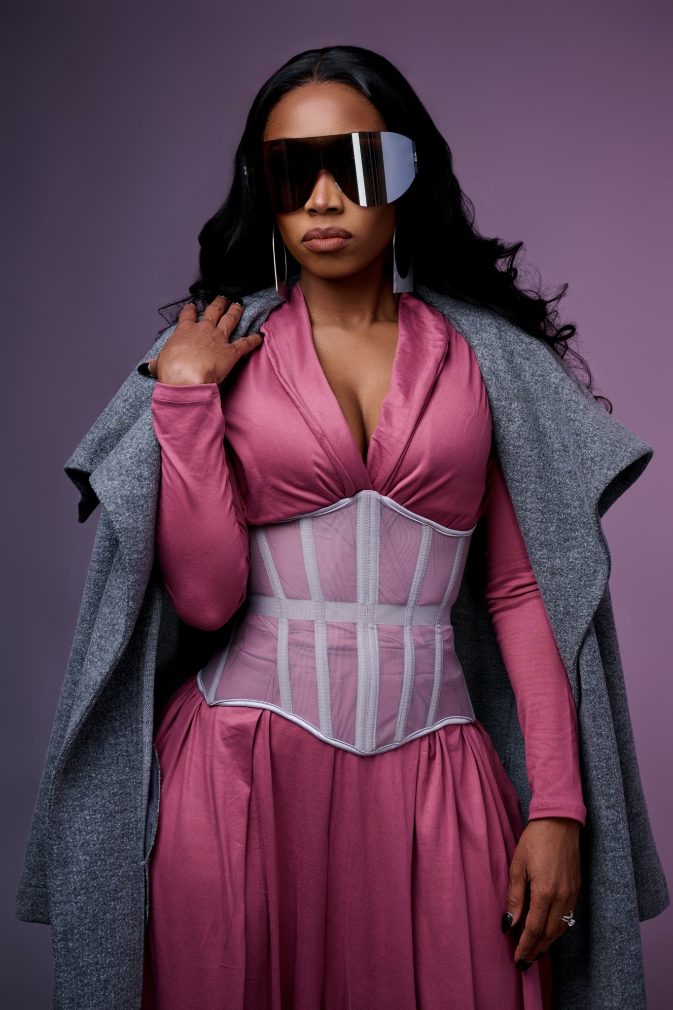 Woman with long dark hair wearing large sunglasses, pink dress, gray coat draped over shoulders, and a waist trainer, standing against purple background.