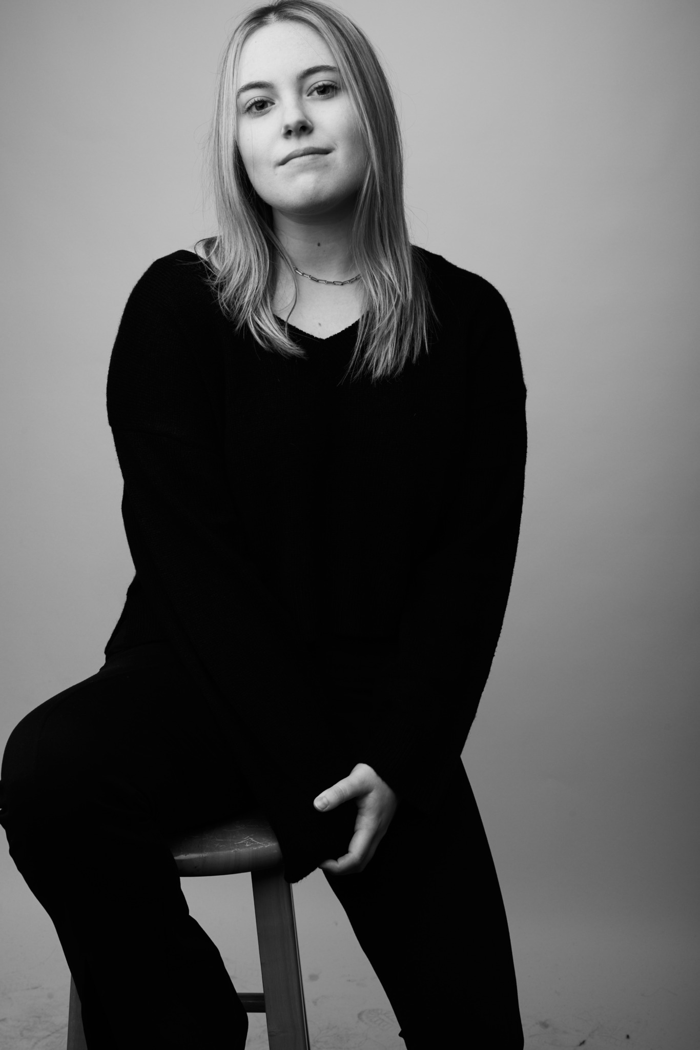 Young woman with long hair sitting on a stool, looking at the camera, wearing a dark top, in black and white.