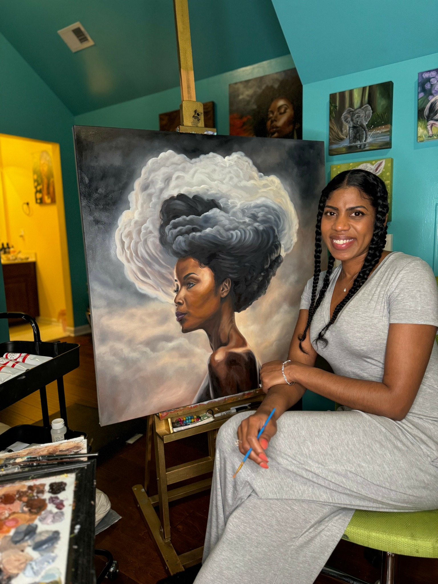 Woman with braided hair sitting next to a painting of a woman with cloud-like hair, in an art studio.