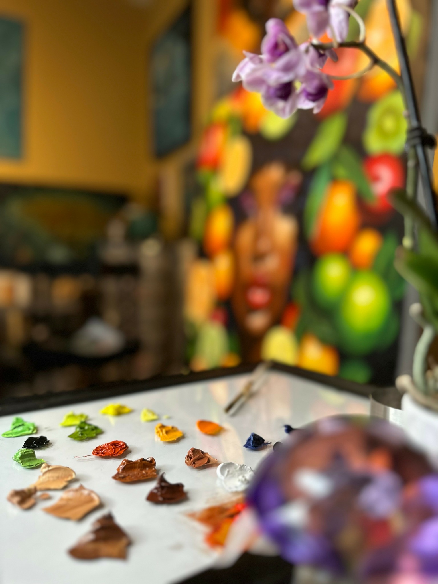 Colorful paint swatches on a white surface with a blurred background of a vibrant, abstract painting and a purple flower.