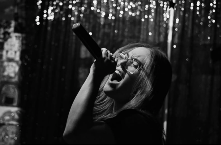 Person singing into a microphone with glasses, in a dark setting with string lights in the background.