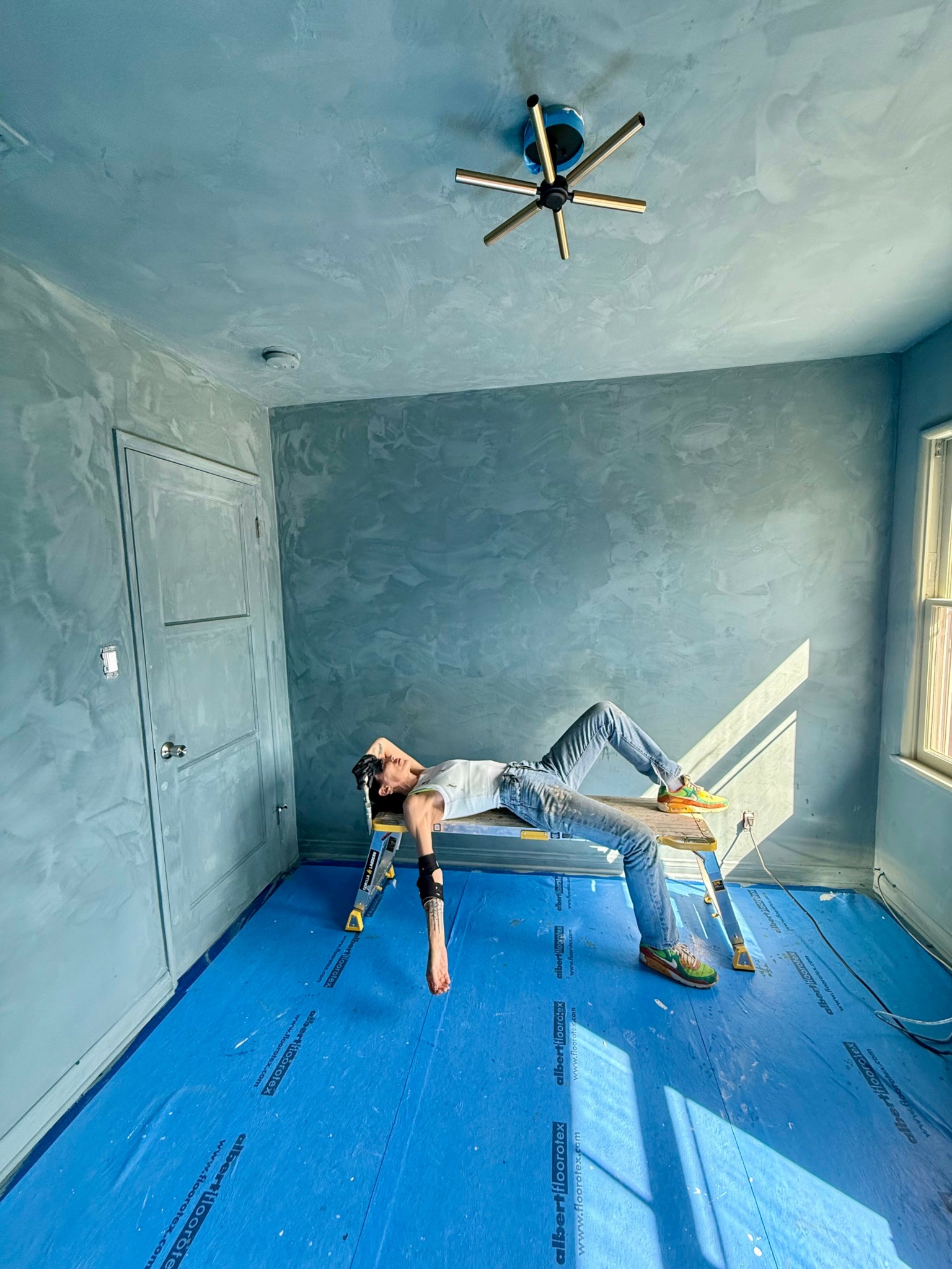 Person lying on a table in a room with blue floor, gray walls, and a ceiling fan, sunlight coming through window.
