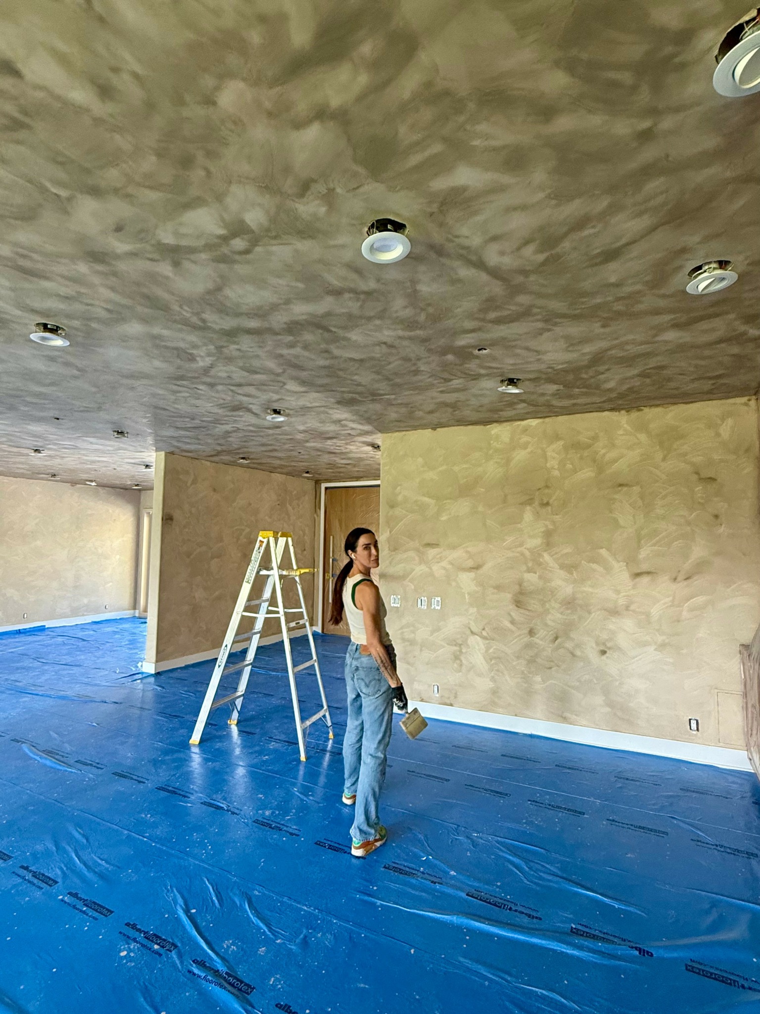 Person standing indoors on blue plastic-covered floor, with unfinished ceiling and beige wall, holding a tool or object.