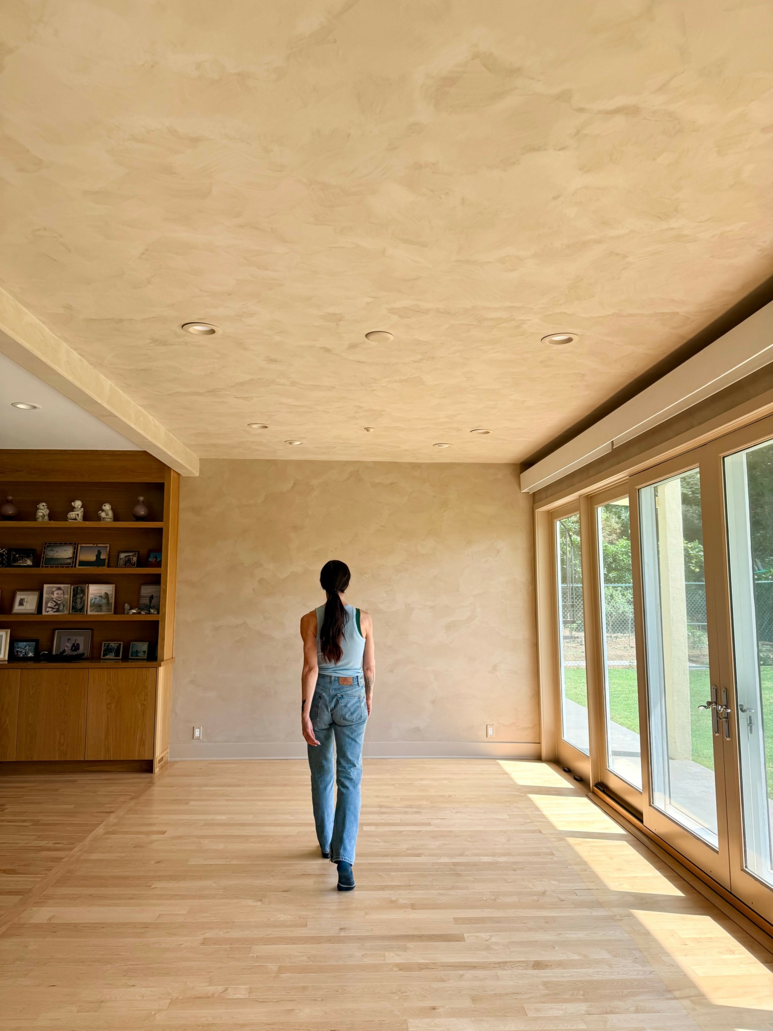Person walking in a spacious room with large windows and wooden flooring, facing away from the camera.