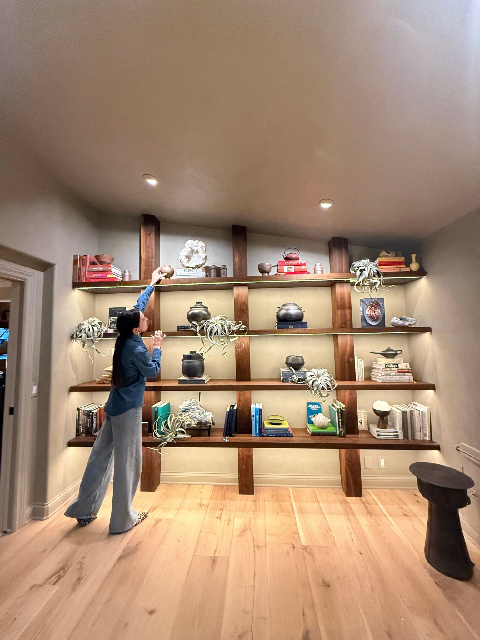 Person standing in front of a wooden bookshelf filled with books and decorative items, in a room with wooden flooring.