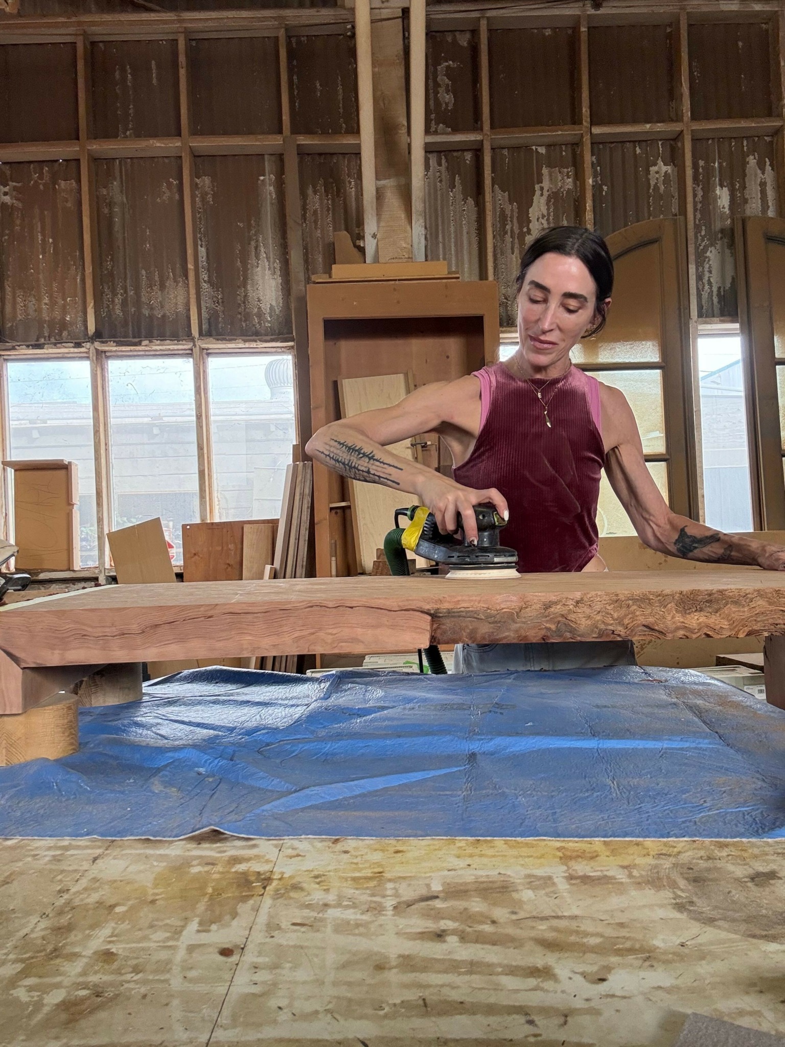 Woman with tattoos working on a wooden project in a workshop, holding a tool, with large windows in the background.
