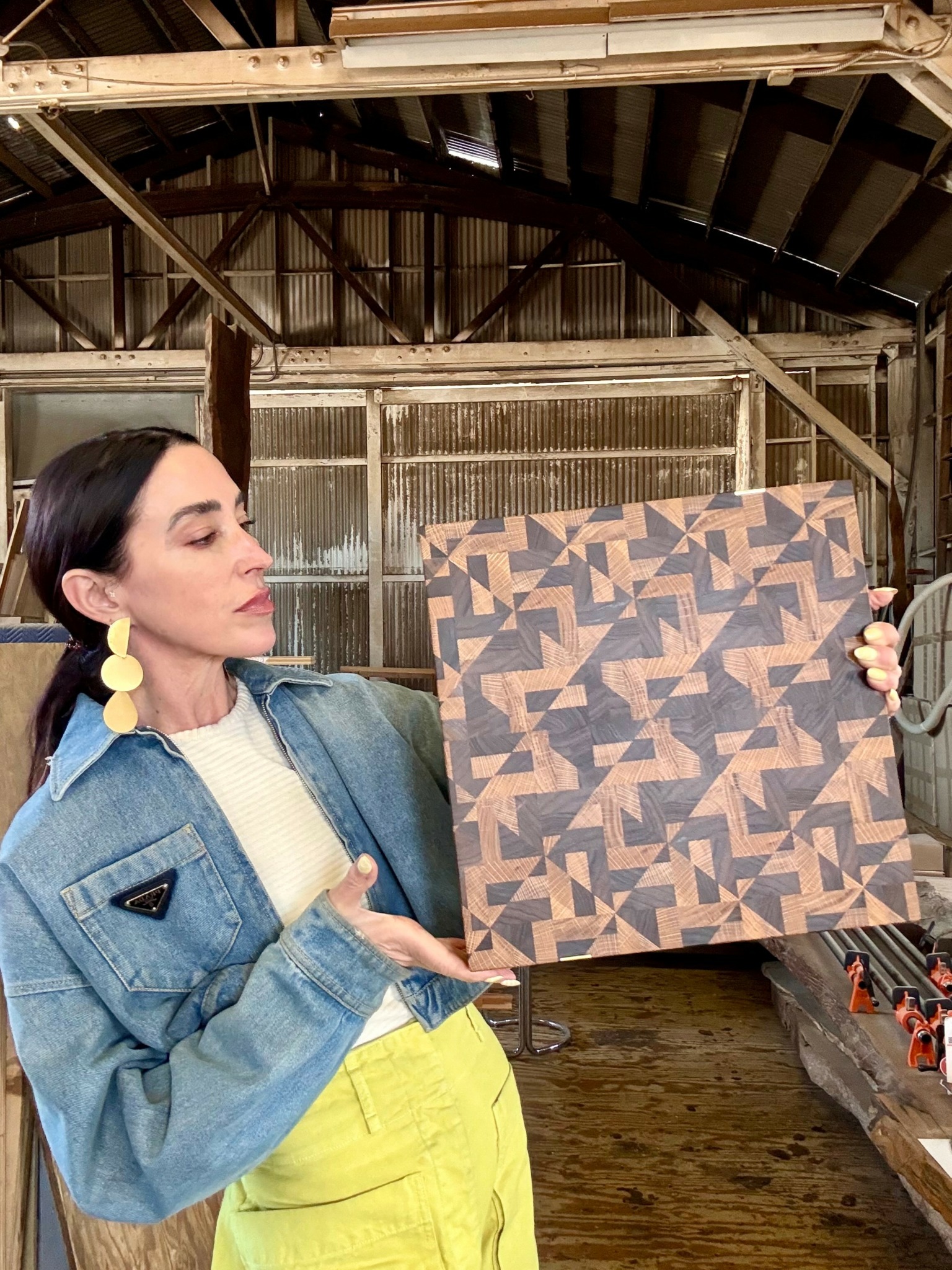 Woman holding a geometric patterned artwork inside a rustic wooden structure.
