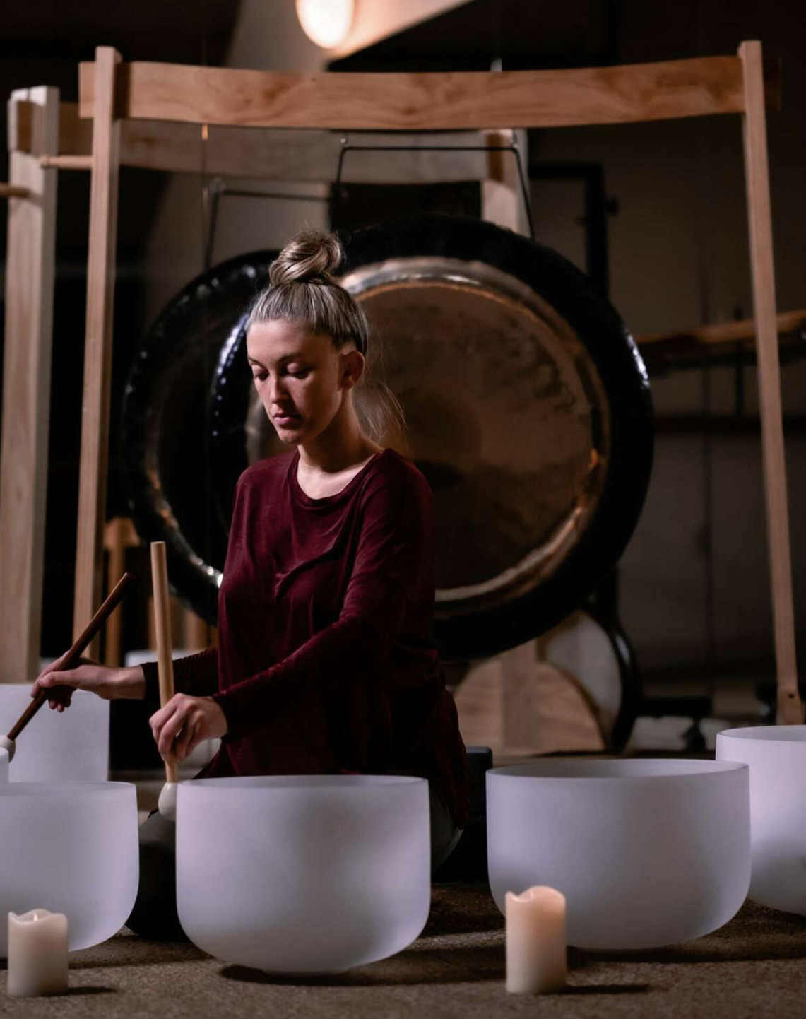 Young woman playing crystal singing bowls in a room with a large gong in the background, candles on the table.