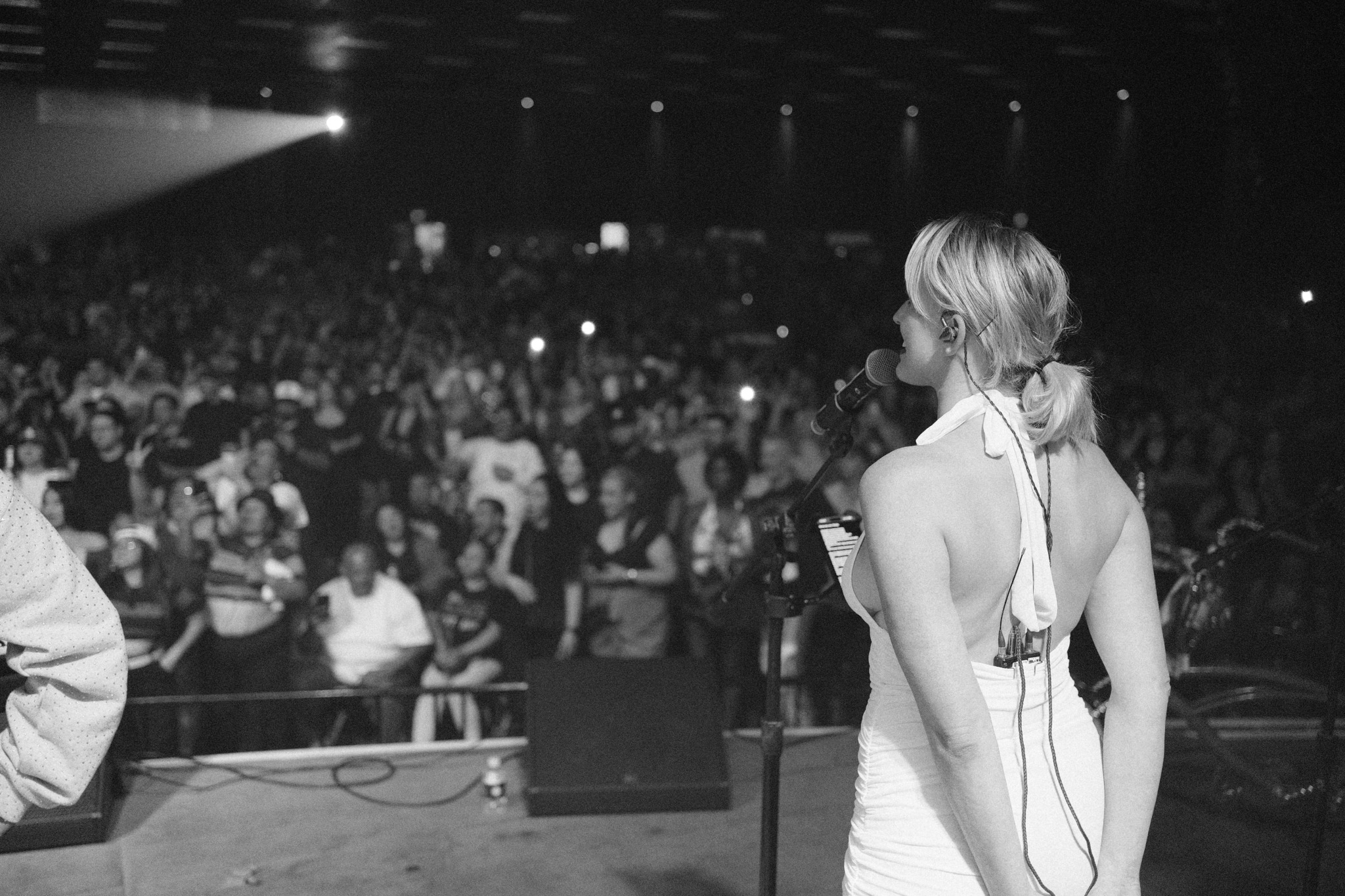 A woman with short blonde hair stands on stage facing a large audience in a dark venue.