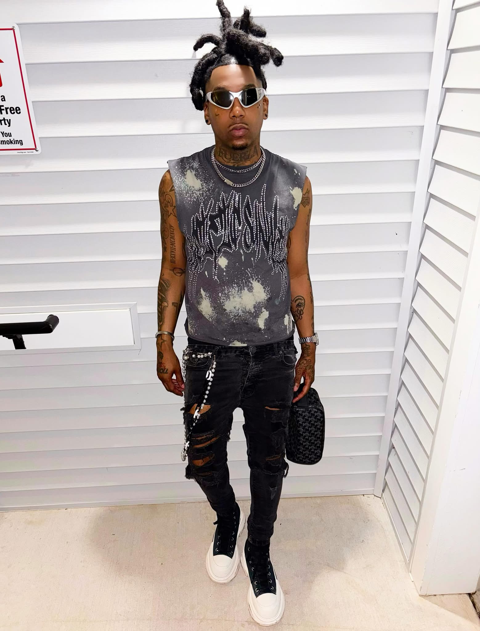 Young man with dreadlocks, sunglasses, sleeveless tie-dye shirt, ripped black jeans, white sneakers, holding a black bag, standing indoors.