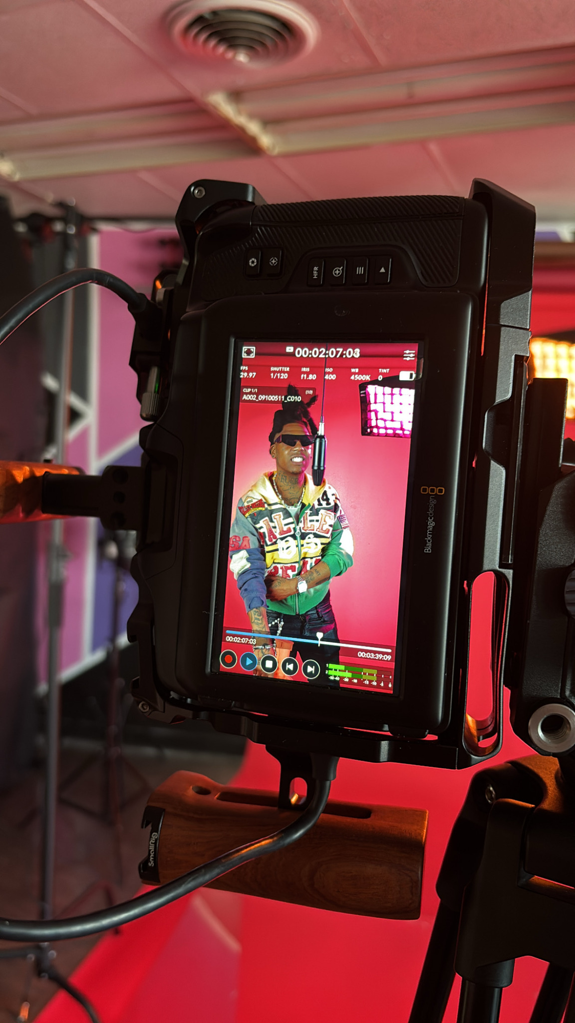 Camera screen shows a person wearing sunglasses and colorful clothing, standing against a pink background.