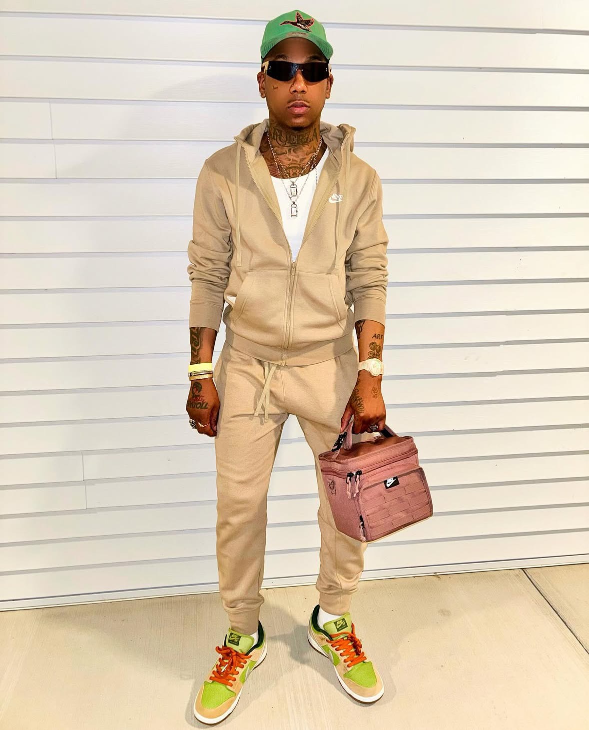 Person in beige tracksuit holding a pink bag, wearing sunglasses and colorful sneakers, standing against a white wall.