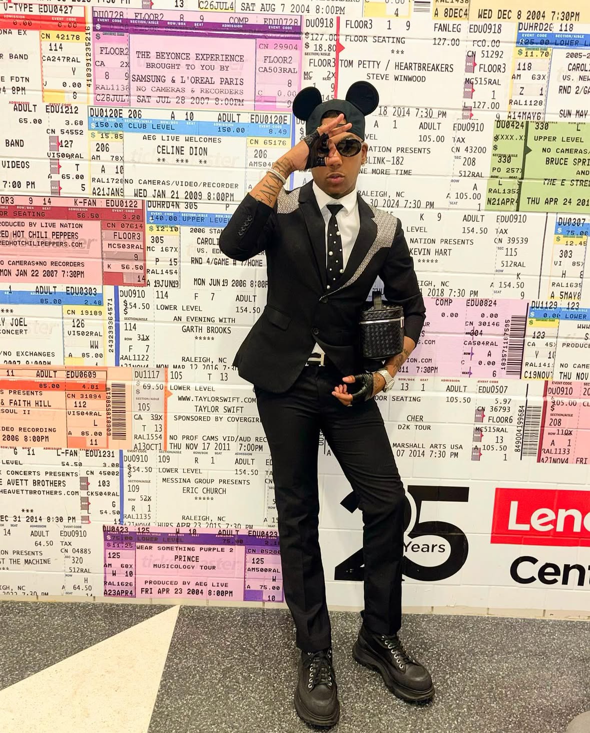 Person in black suit and sunglasses standing in front of a wall covered with colorful travel tickets, holding a clutch bag.