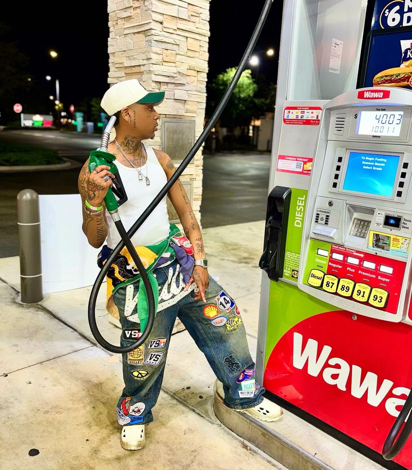 Person at gas station pump holding a green bottle, wearing a white cap, sunglasses, and casual clothing, nighttime scene.