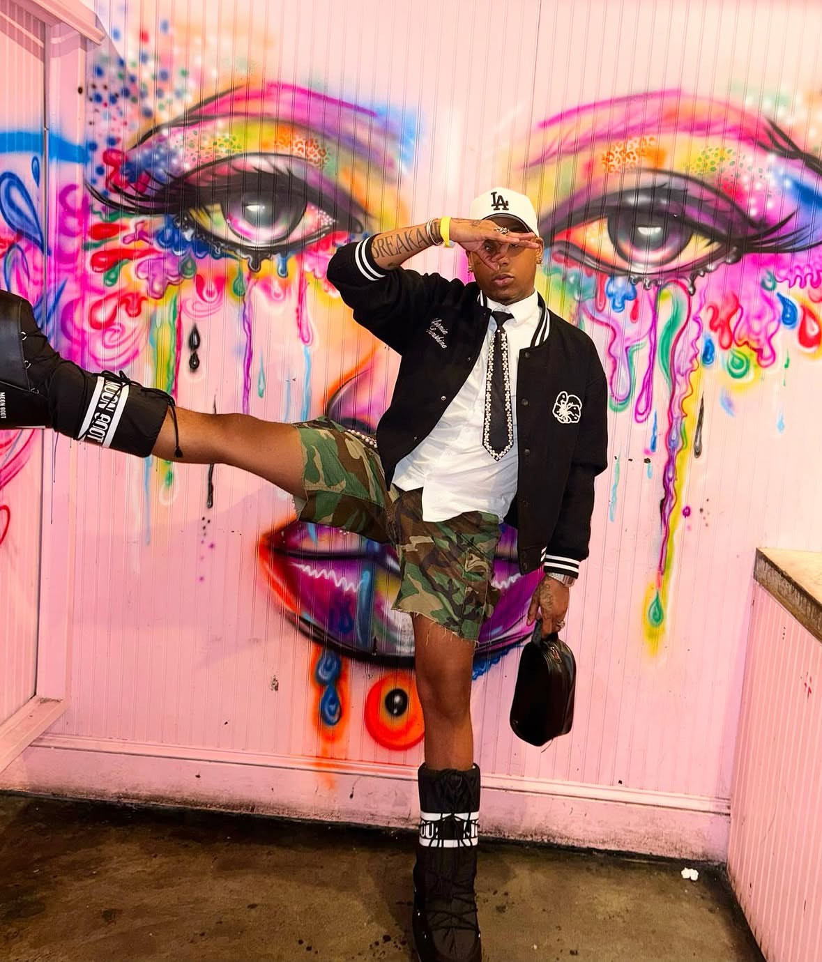 Person standing in front of colorful graffiti mural with eyes and abstract paint drips, wearing a black jacket, white shirt, camouflage shorts, and accessories.
