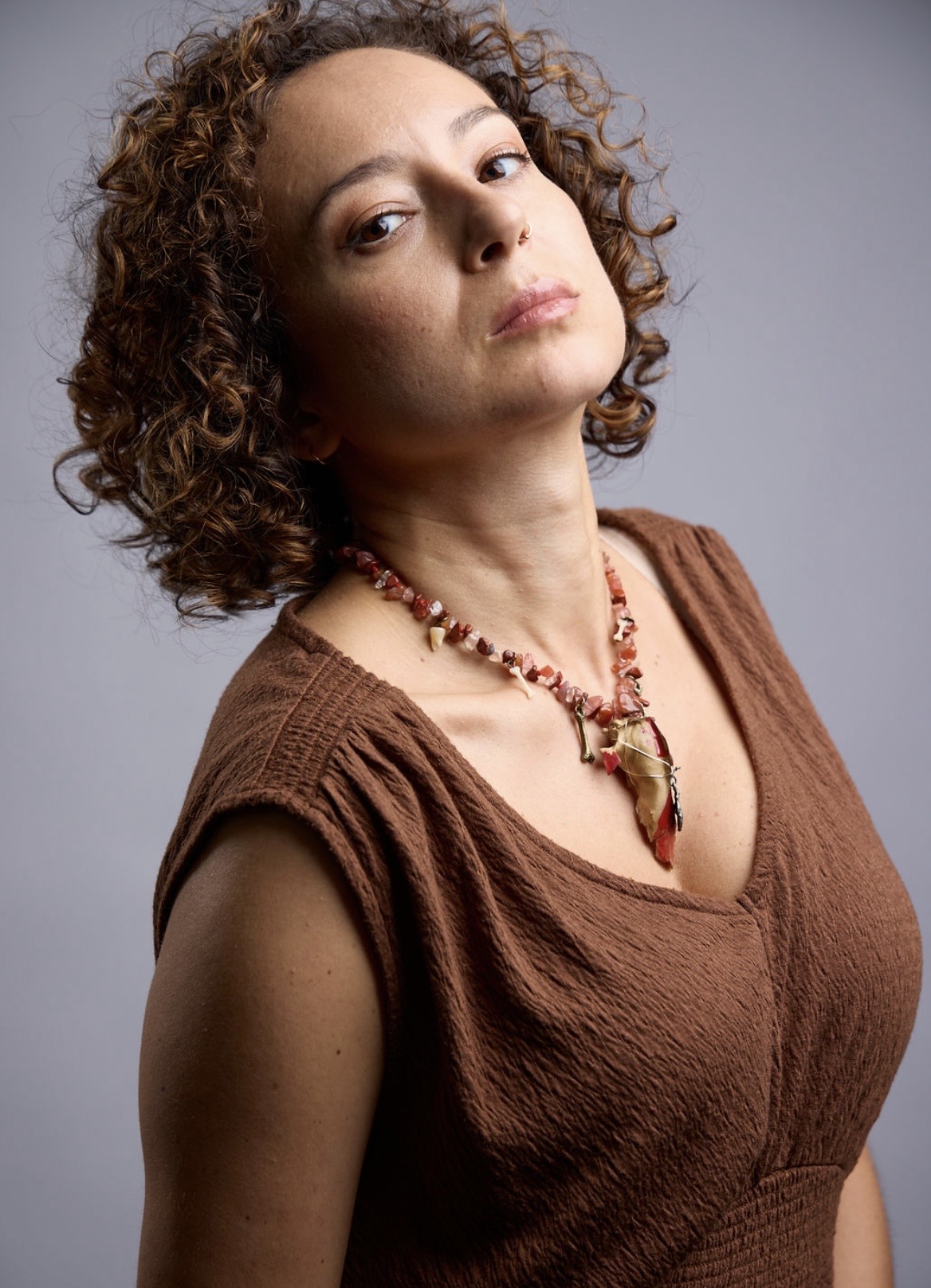 Woman with curly hair wearing a brown top and beaded necklace, looking confidently at the camera.