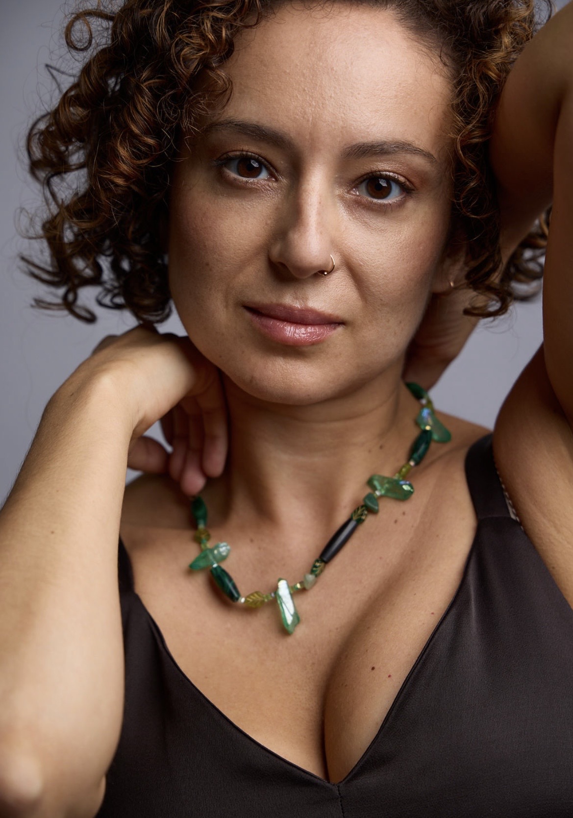 Close-up of a woman with curly hair wearing a black top and a green beaded necklace, looking at the camera.